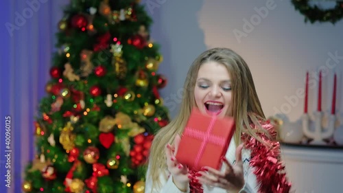 Caucasian woman in a light sweater, slow-motion, jumping with pleasure with a New Year's gift into a cozy decorated room near the Christmas tree. Getting ready for the New Year holiday.
