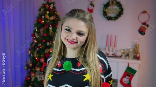 A cheerful cute girl looks at the camera in slow motion 120 fps against the background of a Christmas tree in a New Year's sweater.