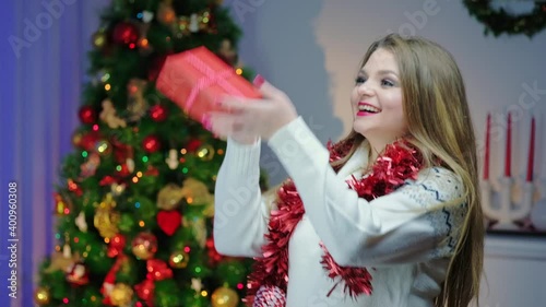Caucasian woman in a light sweater, slow-motion, jumping with pleasure with a New Year's gift into a cozy decorated room near the Christmas tree. Getting ready for the New Year holiday.