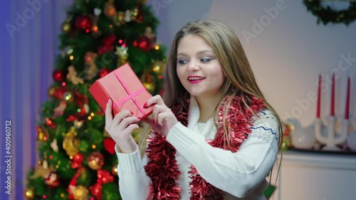Caucasian woman in a light sweater, slow motion shakes a Christmas present in a cozy decorated room near a Christmas tree. Getting ready for the New Year holiday.