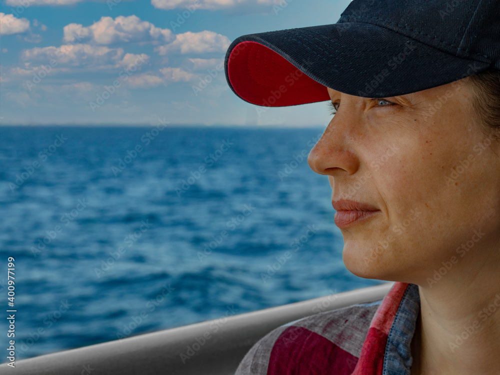 A woman in a baseball cap stands on the deck of a ship and looks into the distance. Nice summer weather. The sea is calm. There are small clouds in the sky.