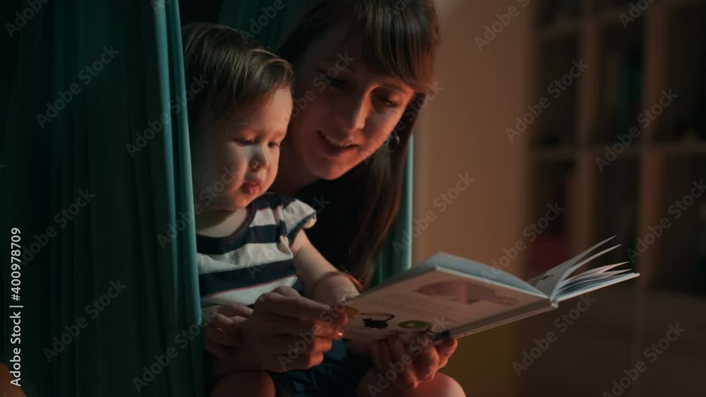 Baby and mother read a hammock. Mother reads a book with together girl being in the hammock at home Baby and mother read a hammock. Mother reads a book with together girl being in the hammock at home