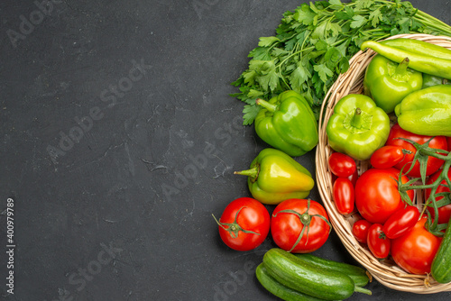 Horizontal view of fresh vegetables red tomatoes with stems green peppers and cucumbers inside and outside of basket on dark background