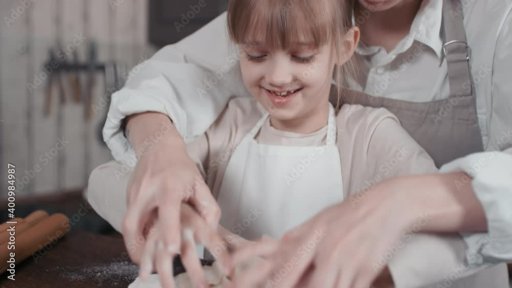Tilting-up medium close-up of smiling caucasian woman teaching little girl knead bread dough with hands standing at kitchen table having fun together Tilting-up medium close-up of smiling caucasian woman teaching little girl knead bread dough with hands standing at kitchen table having fun together