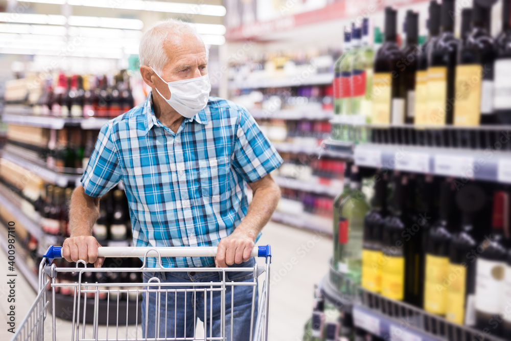 mature european man wearing mask with covid protection chooses bottle of alcohol in supermarket