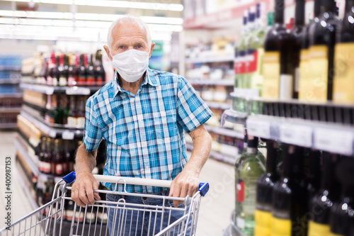 mature european man wearing mask with covid protection chooses bottle of alcohol in supermarket