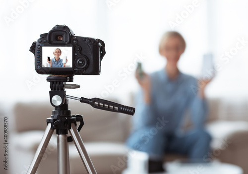 Young Caucasian woman recording video content for her tech blog at home, selective focus on camera with preview screen photo