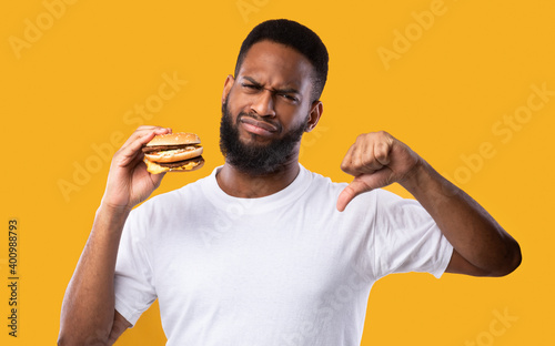 Black Man With Burger Gesturing Thumbs-Down Standing On Yellow Background