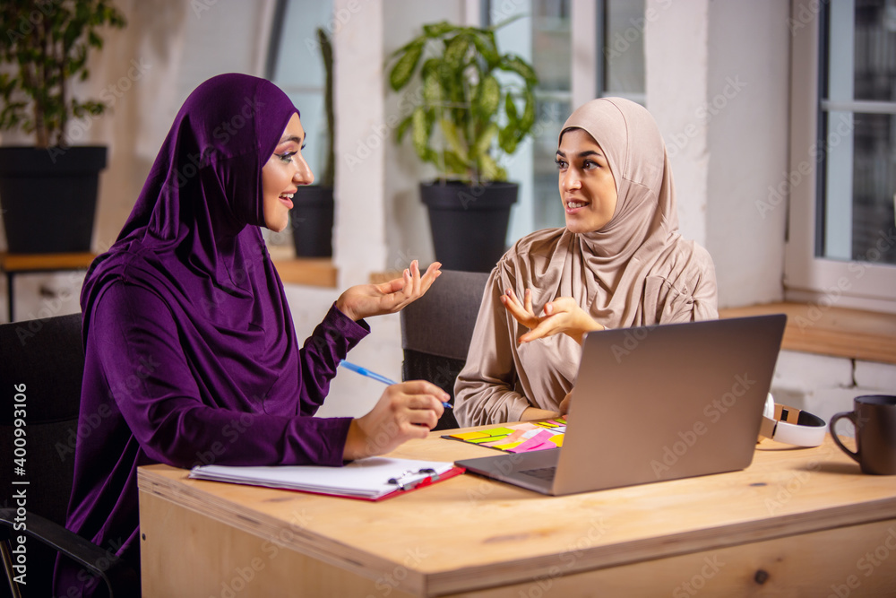 Meanings. Happy and young two muslim women at home during lesson, studying near computer, online education. Culture, traditions, modern people. Watching at PC monitor, shopping or talking. Meanings. Happy and young two muslim women at home during lesson, studying near computer, online education. Culture, traditions, modern people. Watching at PC monitor, shopping or talking.