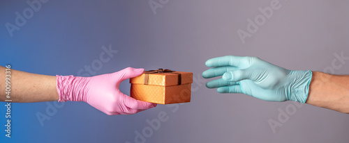 A man's hand in a blue glove is giving a gift box to a woman's hand in pink glove on a blue and gray background. Concept of a lifestyle in a pandemic