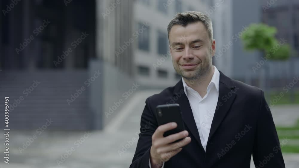 Crop view of handsome mature guy in classical suit and shirt shaking head and looking at smartphone screen. Successful businessman in 40s having video call while walking at street. Crop view of handsome mature guy in classical suit and shirt shaking head and looking at smartphone screen. Successful businessman in 40s having video call while walking at street.