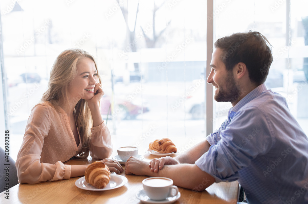 Positive young couple having breakfast together at cafe, sitting near window and enjoying conversation - obrazy, fototapety, plakaty 
