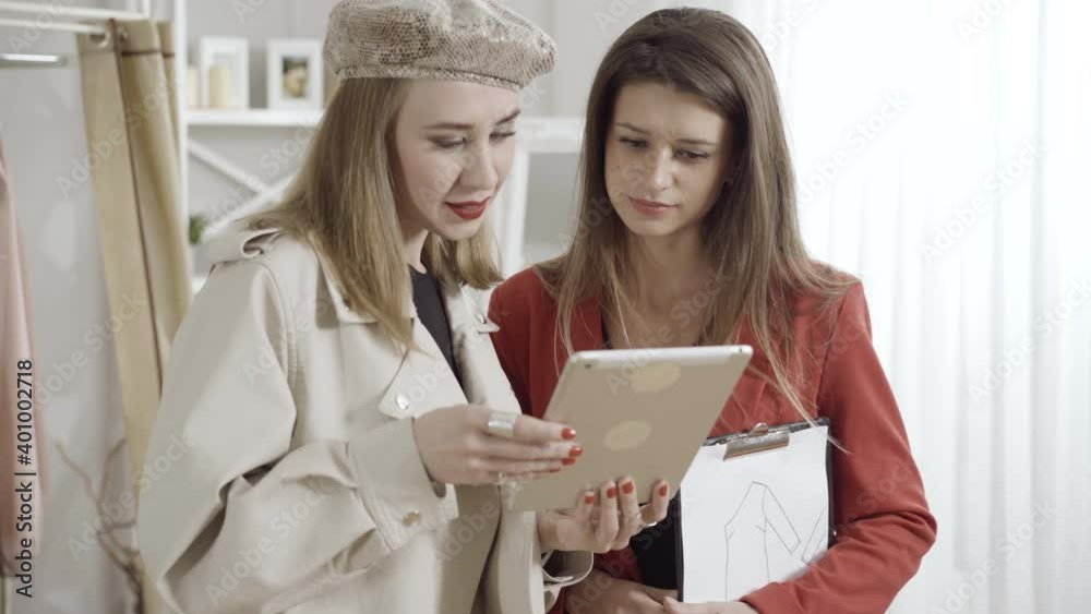 Two positive beautiful clothes designers using tablet and talking indoors. Portrait of charming Caucasian young women discussing fashion design in atelier or workshop. Creativity concept. Two positive beautiful clothes designers using tablet and talking indoors. Portrait of charming Caucasian young women discussing fashion design in atelier or workshop. Creativity concept.