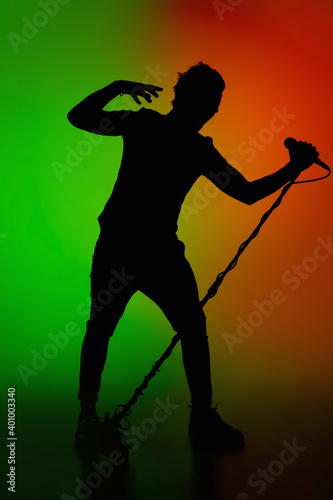 Move. Silhouette of young male guitarist isolated on green-orange gradient studio background in neon. Beautiful shadow in action, performing. Concept of human emotions, expression, ad, music, art.