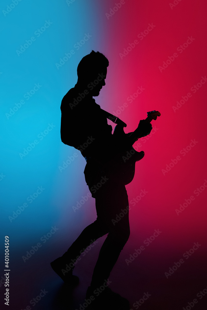 Action. Silhouette of young male guitarist isolated on blue-pink gradient studio background in neon. Beautiful shadow in action, performing. Concept of human emotions, expression, ad, music, art.