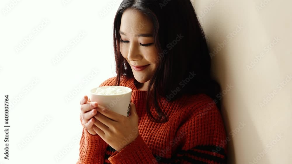 Young beautiful Asian woman holding cup of coffee and marshmallows on top, sitting at home and looking out the window. Happy girl drinking chocolate in sweater in cold weather winter Young beautiful Asian woman holding cup of coffee and marshmallows on top, sitting at home and looking out the window. Happy girl drinking chocolate in sweater in cold weather winter