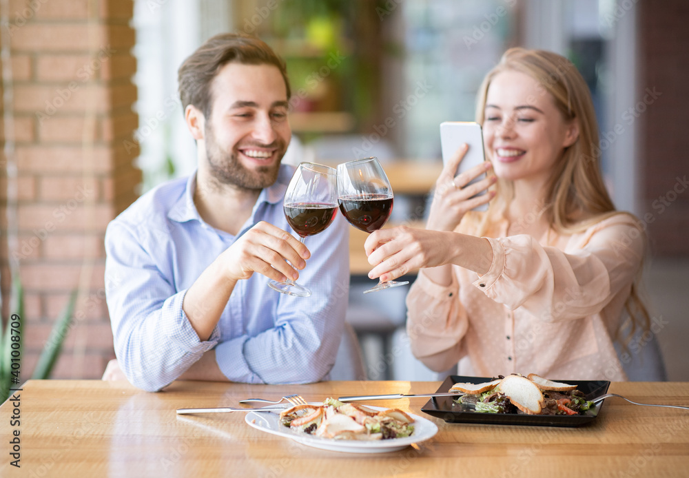Portrait of lovely lady taking picture of toast with clinking wine glasses during holiday dinner at restaurant