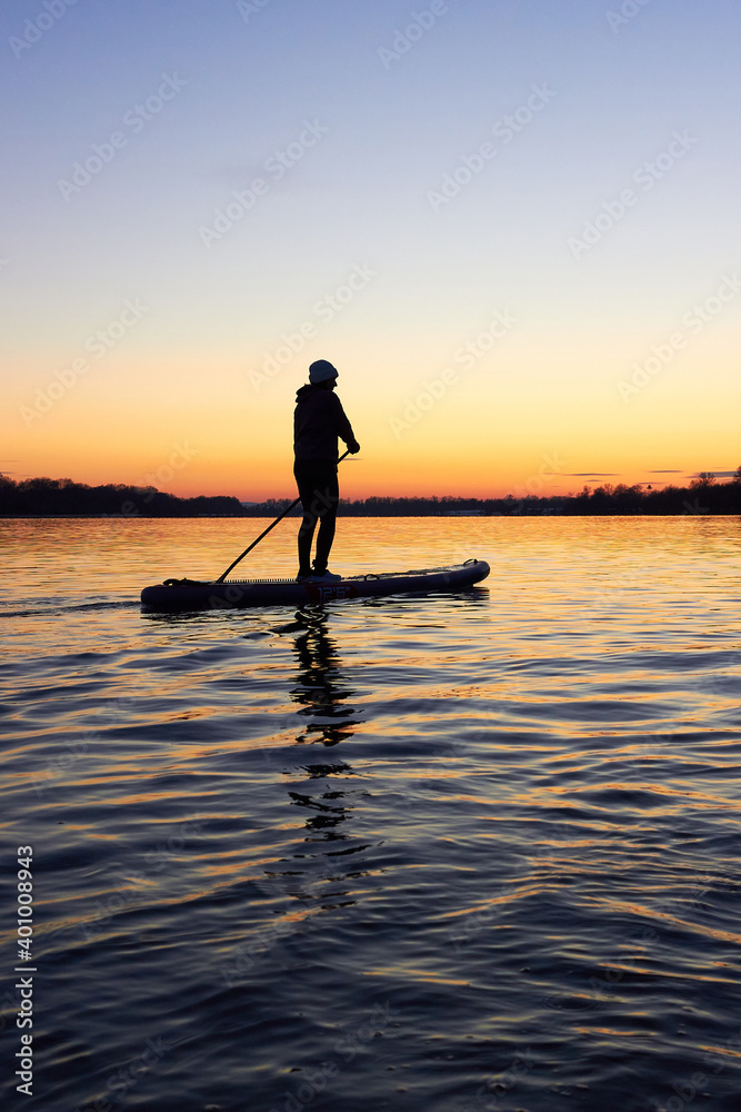 A middle-aged woman on stand up paddle boarding at dusk on a flat quiet winter river with beautiful sunset colors