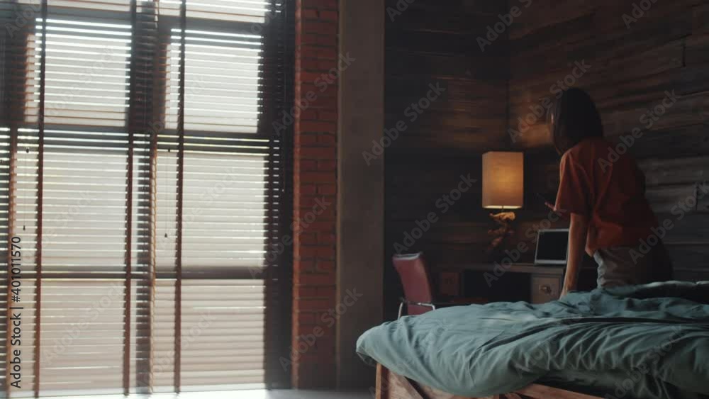 PAN shot of young Asian woman getting out of bed, walking to large window with blinds and messaging on smartphone in the morning at home PAN shot of young Asian woman getting out of bed, walking to large window with blinds and messaging on smartphone in the morning at home