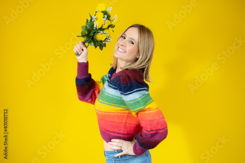 Portrait of a beautiful young woman in colorful sweater holding big bouquet of irises and tulips isolated over yellow background