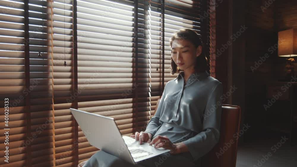 Zoom in shot of young beautiful Asian woman sitting near window with blinds and browsing the Internet on laptop while working in home office