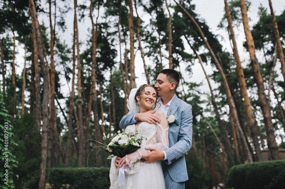 Stylish groom in a blue suit and a beautiful bride in a white lace dress tenderly hug in the forest, in nature with tall pines. Wedding portrait of the newlyweds. Stylish groom in a blue suit and a beautiful bride in a white lace dress tenderly hug in the forest, in nature with tall pines. Wedding portrait of the newlyweds.