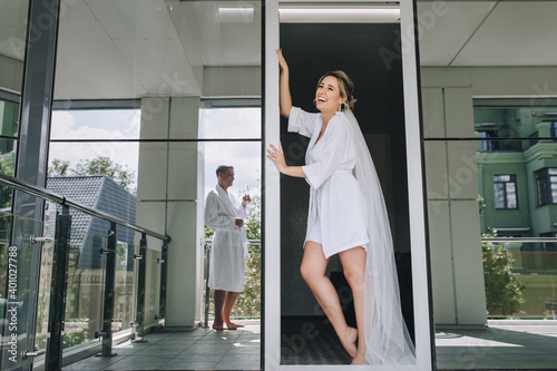 A beautiful and sexy bride, a model in a white robe and a long veil, stands in the doorway against the background of a mirrored wall and looks at the groom with a glass of champagne.