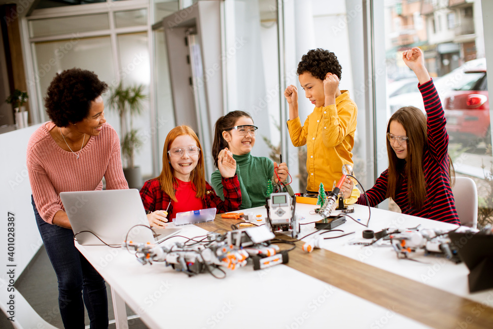 Happy kids with their African American female science teacher with laptop programming electric toys and robots at robotics classroom Happy kids with their African American female science teacher with laptop programming electric toys and robots at robotics classroom