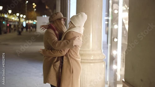 Reconnect of 2 young women, happy and excited to see each other again, embrace each other with enthusiasm. They are in a city at night and wear winter clothes. Together again