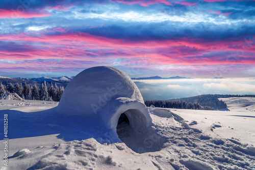 Real snow igloo house in the winter mountains glowing by morning sunlight. Incredible purple sunset with glowing clouds on the background. Winter holiday concept