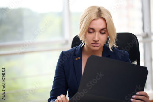 Photo of serious manager or director woman wearing formal clothing and eyeglasses holding in hands paper documents while working in office on laptop