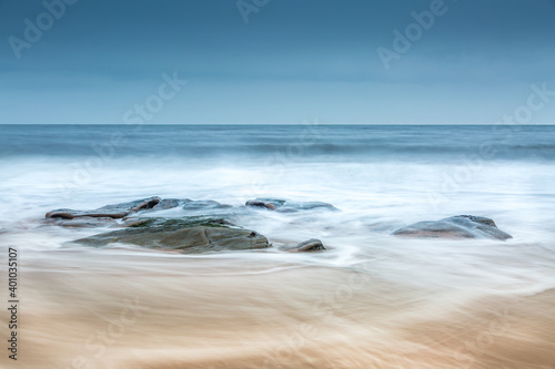 A group of rocks on the beach as the wave backwash returns to the sea over the sand on a cloudy day (2)