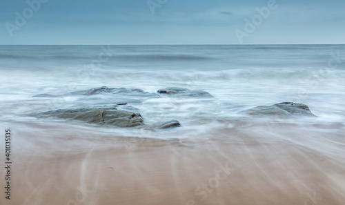 A group of rocks on the beach as the wave backwash returns to the sea over the sand on a cloudy day (1)