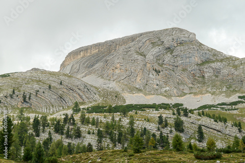 A close up view on a sharp and stony mountain in Italian Dolomites. The massive mountain is surrounded by clouds. A few trees overgrowing the lower parts of it. Remote and desolate area. Overcast photo