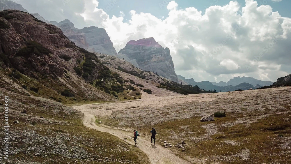 A couple hiking along a gravelled pathway leading through a valley in Italian Dolomites. High, sharp mountains around. Stony and raw landscape. Remote and desolate place. Freedom of exploration