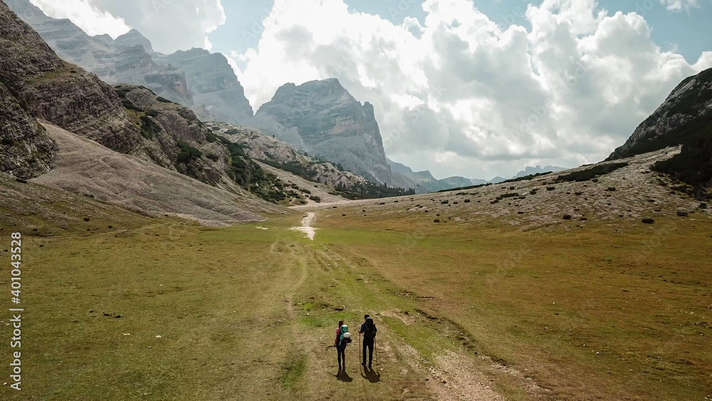 A couple hiking along a pathway leading through a valley in Italian Dolomites. High, sharp mountains around. Stony and raw landscape, green meadow. Remote and desolate place. Freedom of exploration