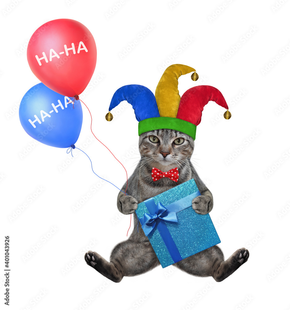 A gray cat clown in a red bow tie and a jester hat is holding a gift box and two balloons. White background. Isolated. A gray cat clown in a red bow tie and a jester hat is holding a gift box and two balloons. White background. Isolated.