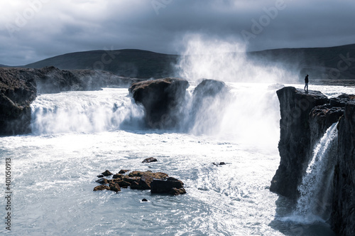 One man standing on a cliff facing the magnificent and tumultuous Goðafoss (waterfall of the gods) in a cool moody light