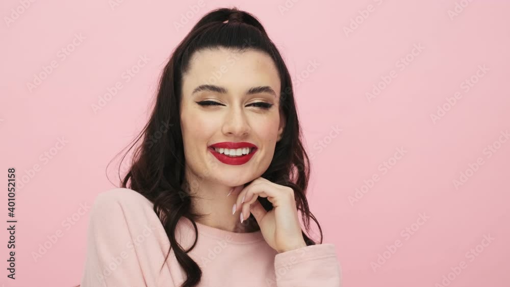 A dreaming woman with red lips is posing to the camera standing isolated over pink background in studio A dreaming woman with red lips is posing to the camera standing isolated over pink background in studio