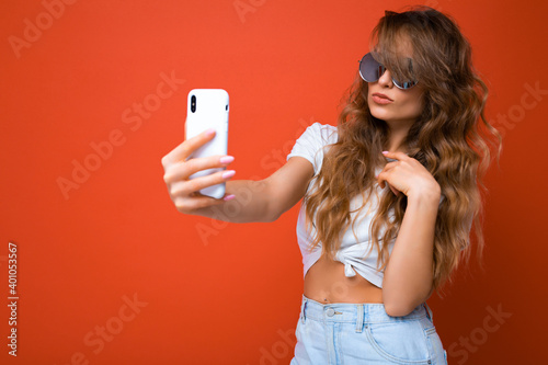 Closeup photo of amazing beautiful young blonde woman holding mobile phone taking selfie photo using smartphone camera wearing sunglasses everyday stylish outfit isolated over colorful wall background