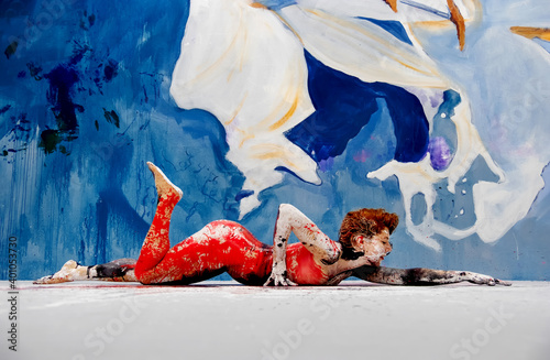 Bodypainting, side view of an artistically painted with red color sexy young woman in the studio with lilies painted wall lying on the floor, copy space
