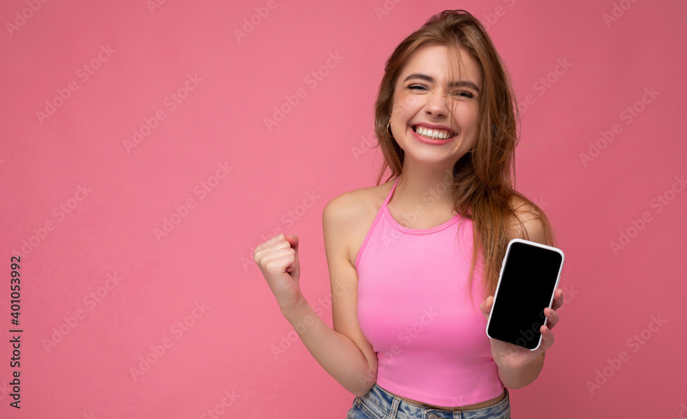 Beautiful joyful young blonde woman wearing pink top poising isolated on pink background with empty space holding in hand and showing mobile phone with empty display for mockup and showing yes gesture Beautiful joyful young blonde woman wearing pink top poising isolated on pink background with empty space holding in hand and showing mobile phone with empty display for mockup and showing yes gesture