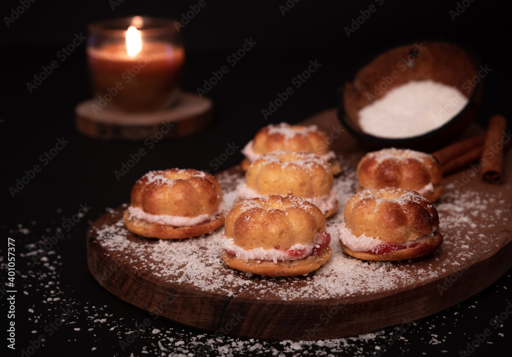 Coconut shell full of flour, sprinkled over five small home baked cakes, filled with strawberry cream. Dark wooden board, cinnamon sticks decoration, burning candle. Black background