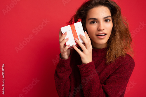 Portrait shot of attractive happy thoughtful amazed adult brunette curly woman isolated over red background wall wearing dark red sweater holding white gift box looking at camera
