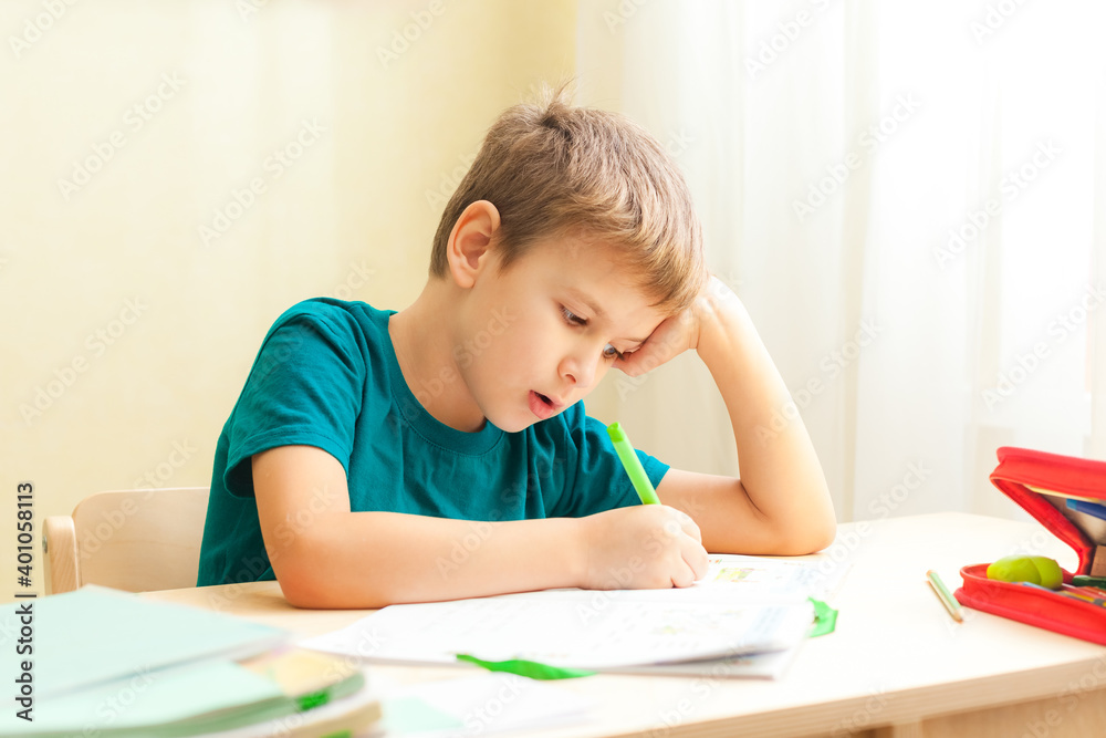 7 yeas old boy sitting at desk and doing homework. Child write notes in notebook, difficult homework, child concentrating on examples 7 yeas old boy sitting at desk and doing homework. Child write notes in notebook, difficult homework, child concentrating on examples