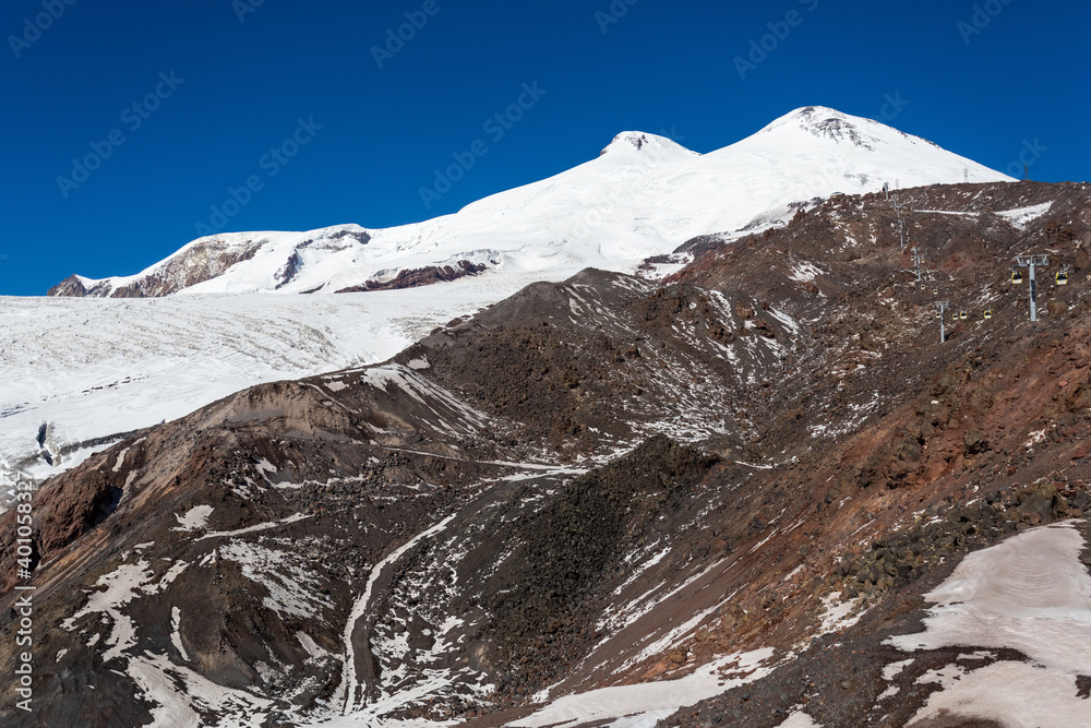 Rocks, white slopes and the summit of Elbrus, Caucasus. Snow-covered peaks and clear blue skies. Winter mountains, alpine skiing. Sunny snowy landscape. Panoramic mountain view.