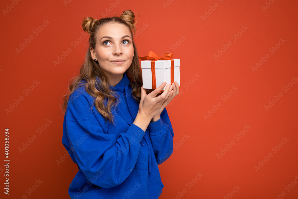 Beautiful happy young blonde woman isolated over colourful background wall wearing stylish casual clothes holding gift box and looking to the side Beautiful happy young blonde woman isolated over colourful background wall wearing stylish casual clothes holding gift box and looking to the side