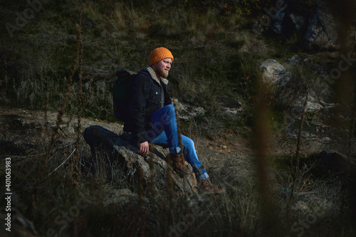 Side view of a lamber man in orange knitted woolen having rest on a rock in the steeppe after hiking and looking ahead. Plants in the foreground. Concept of people traveling in nature. photo