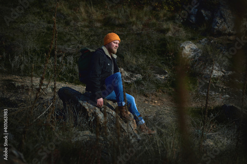 Side view of a lamber man in orange knitted woolen having rest on a rock in the steeppe after hiking and looking ahead. Plants in the foreground. Concept of people traveling in nature.