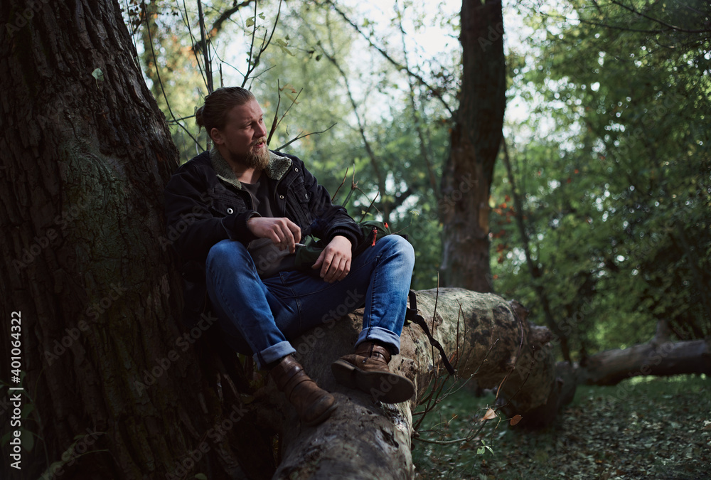 Portrait of a smoking young bearded man adventurer relaxing in the autumn forest and holding a cigarette Portrait of a smoking young bearded man adventurer relaxing in the autumn forest and holding a cigarette